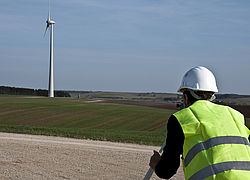 Eine Person im Vordergrund mit Warnweste und Schutzhelm nimmt eine Landvermessung vor. Im Hintergrund flache Landschaft und Windkraftanlage.