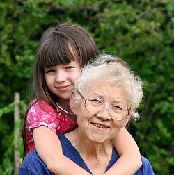 Ein kleines lächelndes Mädchen sitzt Huckepack auf dem Rücken einer alten, lächelnden Frau und umarmt sie