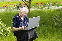 Alte Frau sitzt mit einem Laptop im grünen Garten und tippt